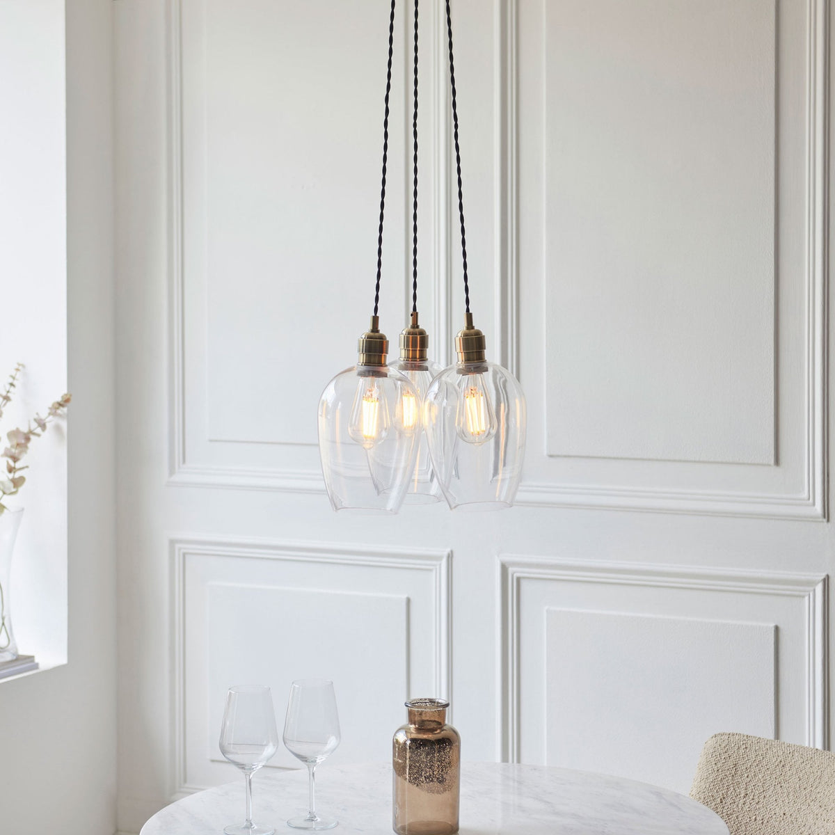 Three clear glass pendant lights with brass fittings hang above a white marble table in a room with wainscoting.