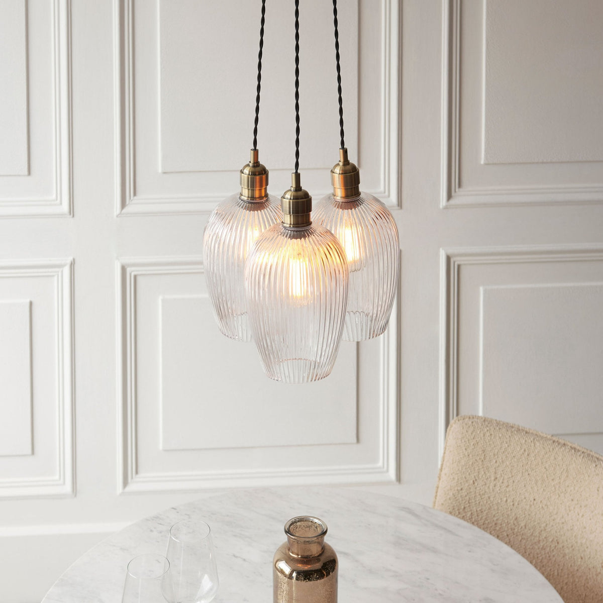 Three ribbed glass pendant lights with brass fittings hang above a marble table with a bronze vase.