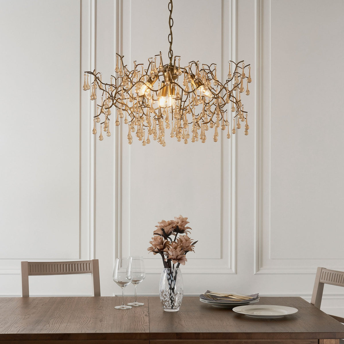 A bronze 12-light chandelier hanging above a dining table set with plates, glasses, and a vase of flowers.