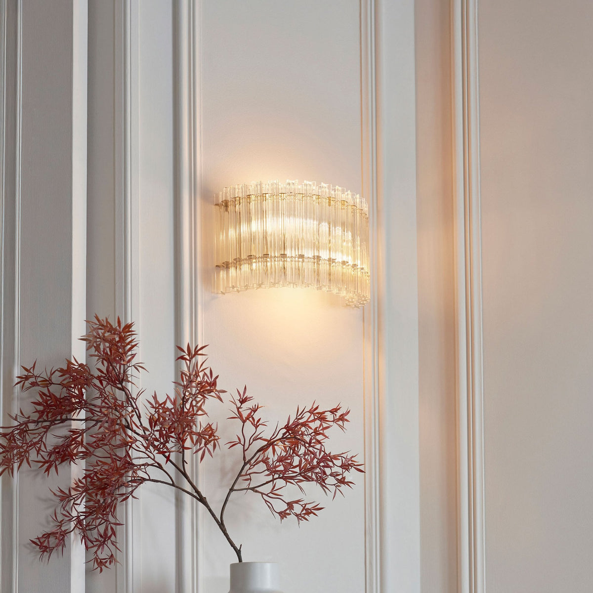 A curved glass and brass wall sconce with vertical fluted glass rods illuminating a white paneled wall above red foliage.