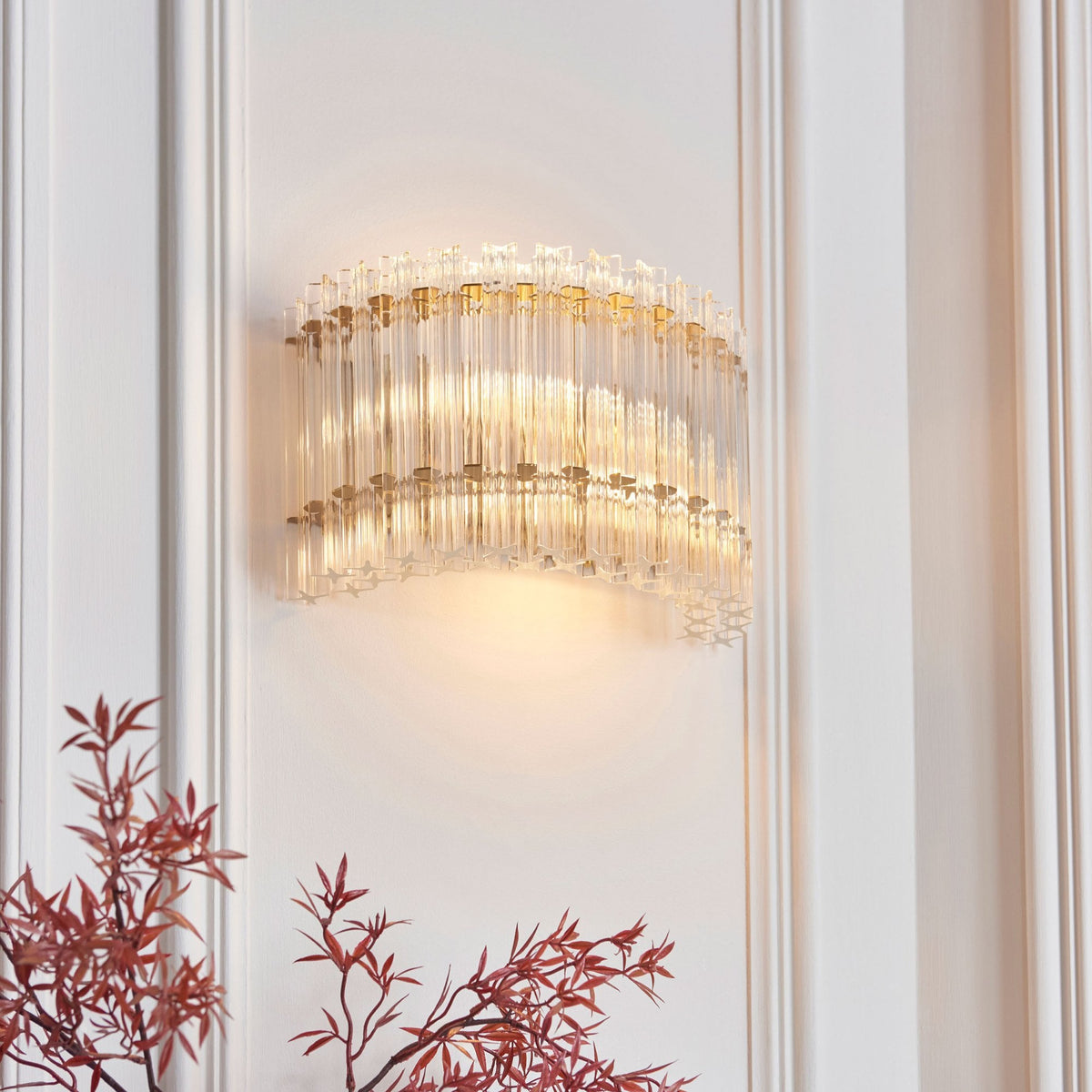 A curved glass wall sconce with a warm glow on a white paneled wall, with a decorative red plant below.