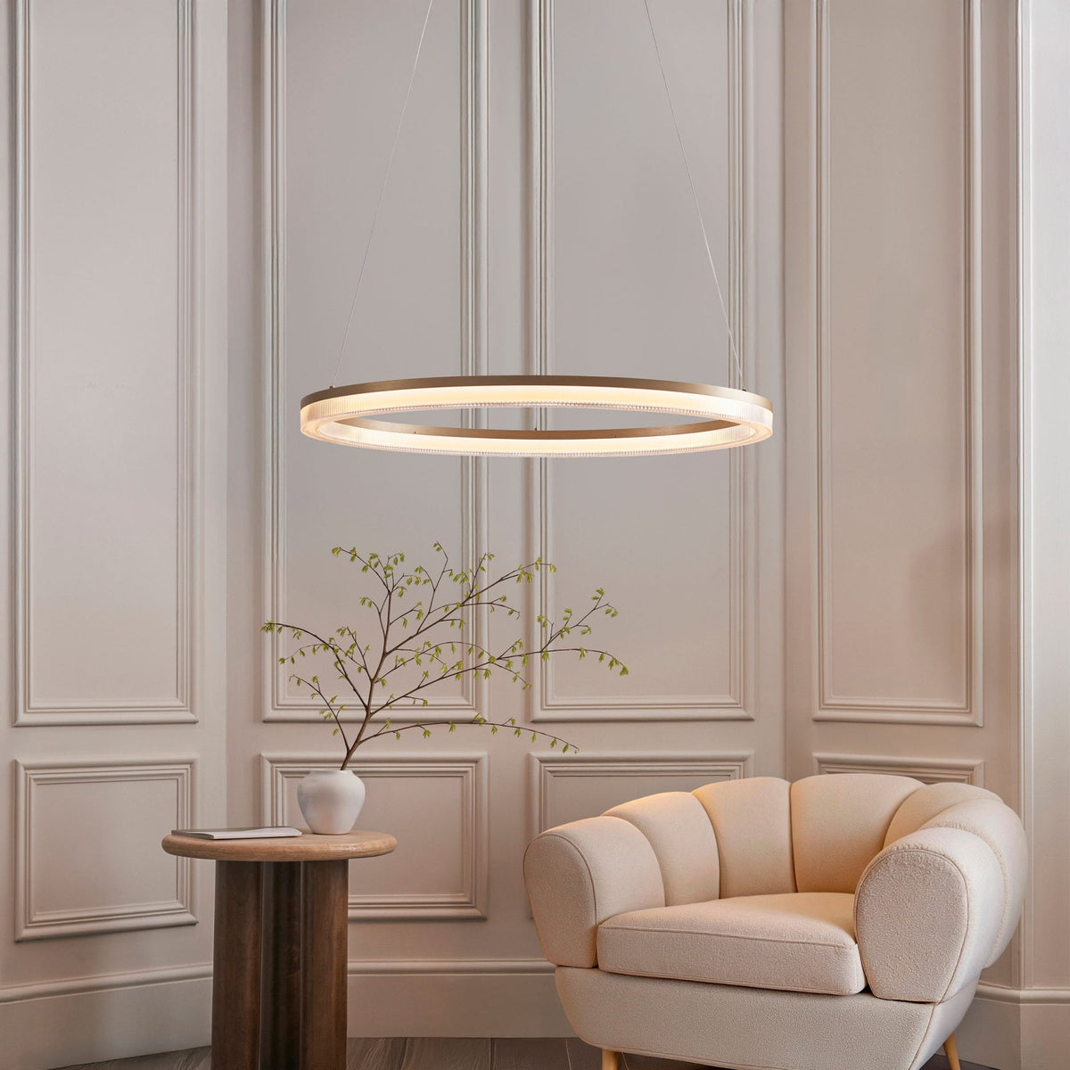 An illuminated gold ring-shaped chandelier hangs above a side table and a beige armchair in a room.