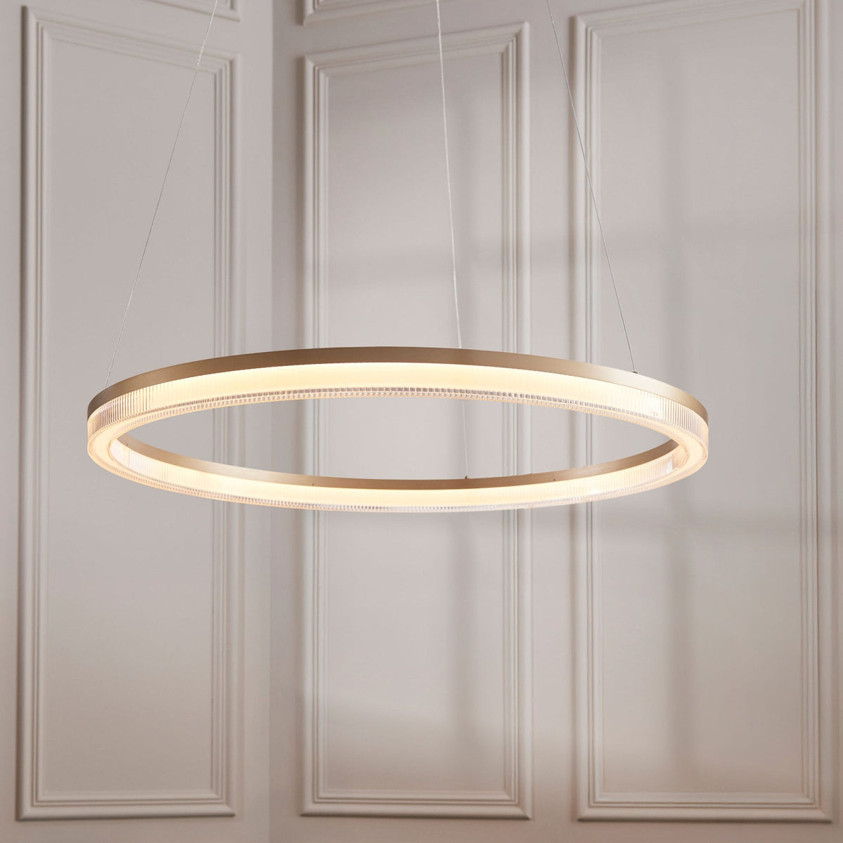 An illuminated gold ring-shaped chandelier hangs from a ceiling against a wall with decorative paneling.
