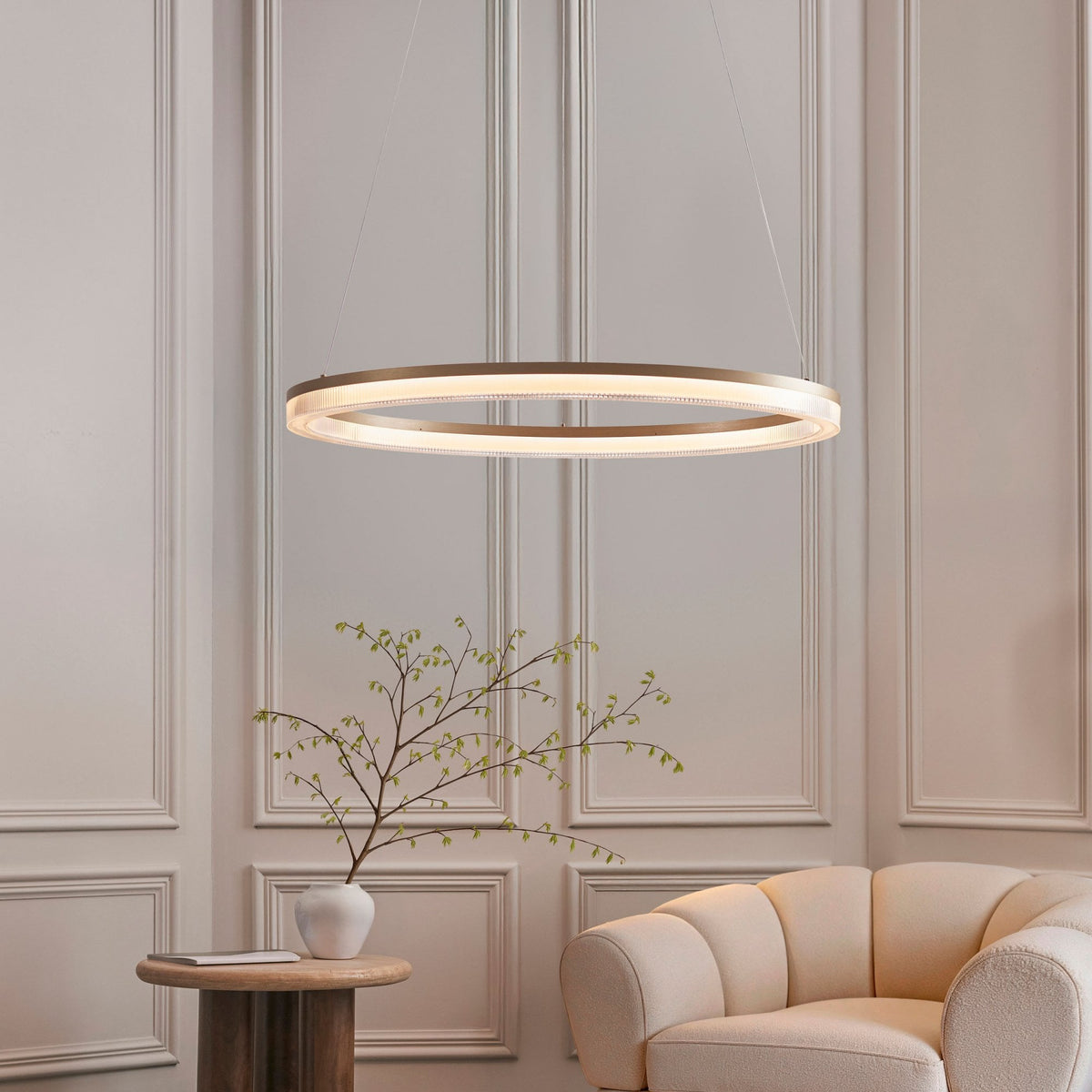 An illuminated gold ring chandelier elegantly hangs above a side table with a plant and a beige armchair.