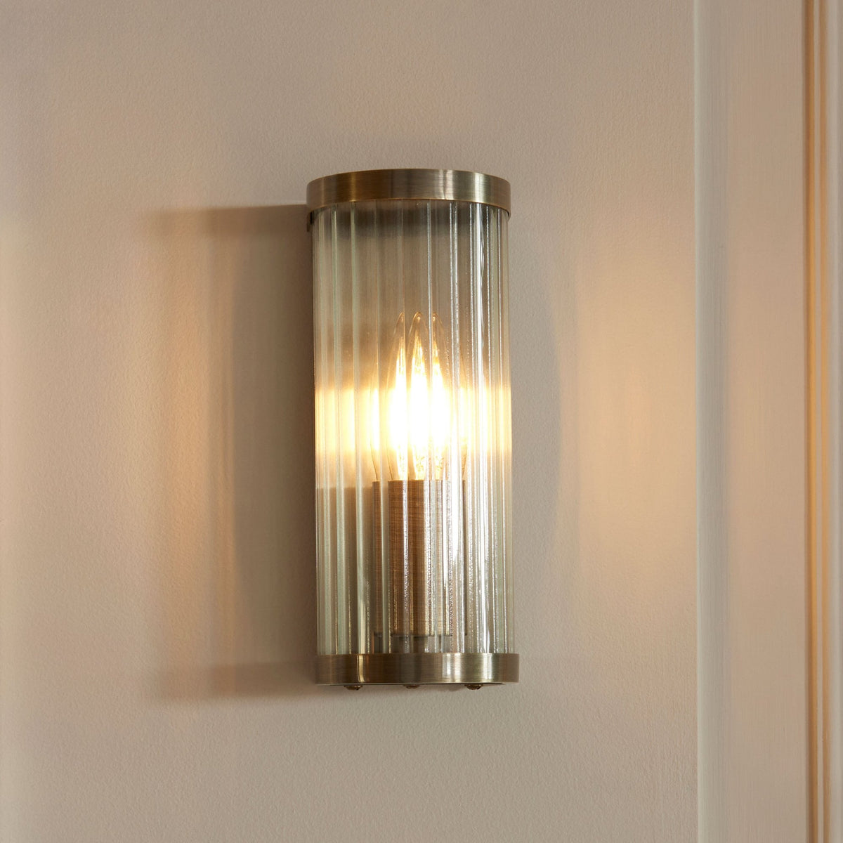 A sleek, gold-plated, wall-mounted light with a fluted glass shade, glowing warmly on a white wall.