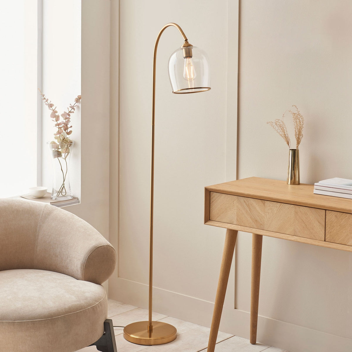 A brass floor lamp with a clear glass shade next to a beige armchair and a wooden desk.