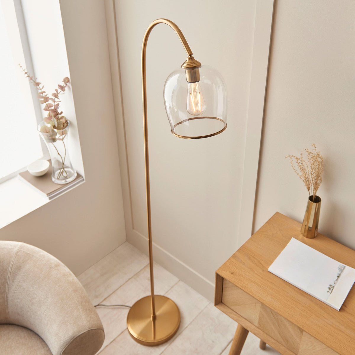 A brass floor lamp with a clear glass shade stands beside a light-colored armchair and a wooden table.
