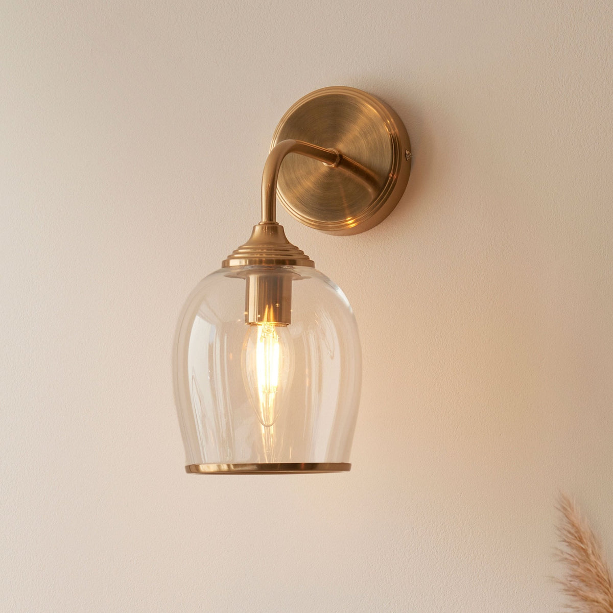 A close-up shot of a brass wall light with a clear glass bell-shaped shade, illuminated, casting a warm glow.