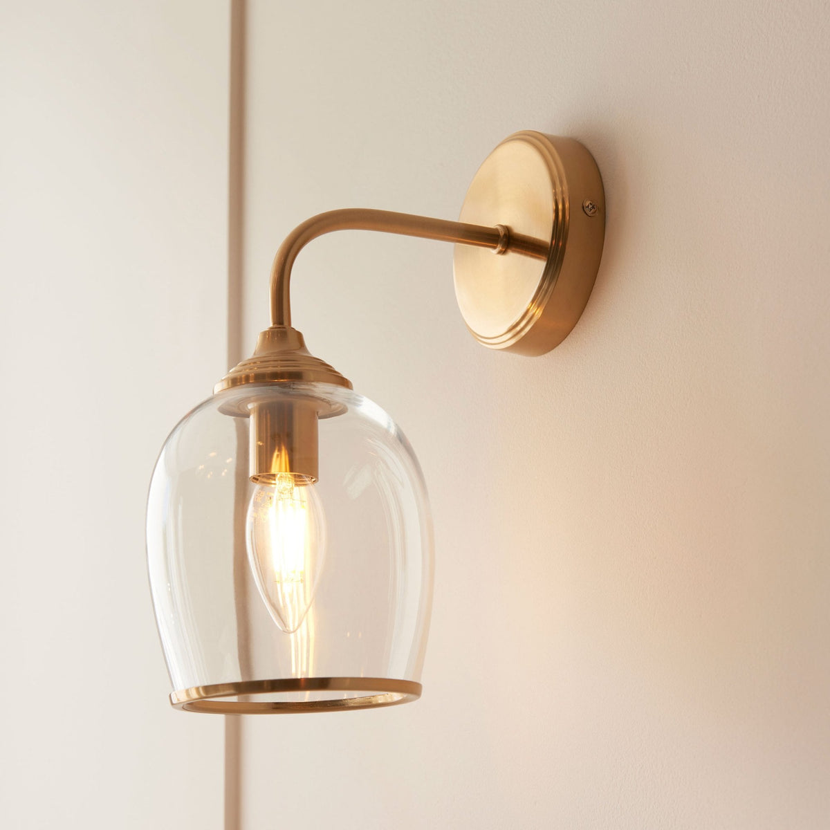 A brass wall light with a curved arm and a clear glass shade, providing ambient lighting against a light wall.