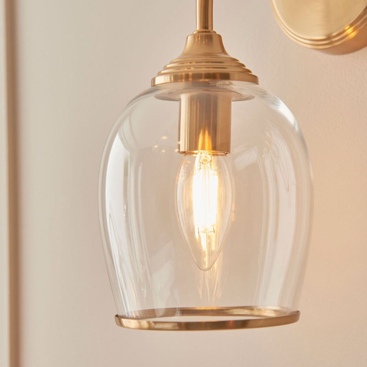 A close-up of the clear glass bell shade and the illuminated filament bulb of the brass wall light.