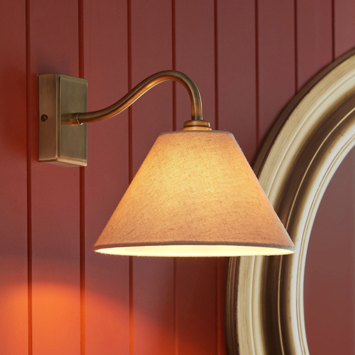 Aplique de pared de latón antiguo con pantalla cónica gris, emitiendo luz cálida sobre una pared de madera roja.