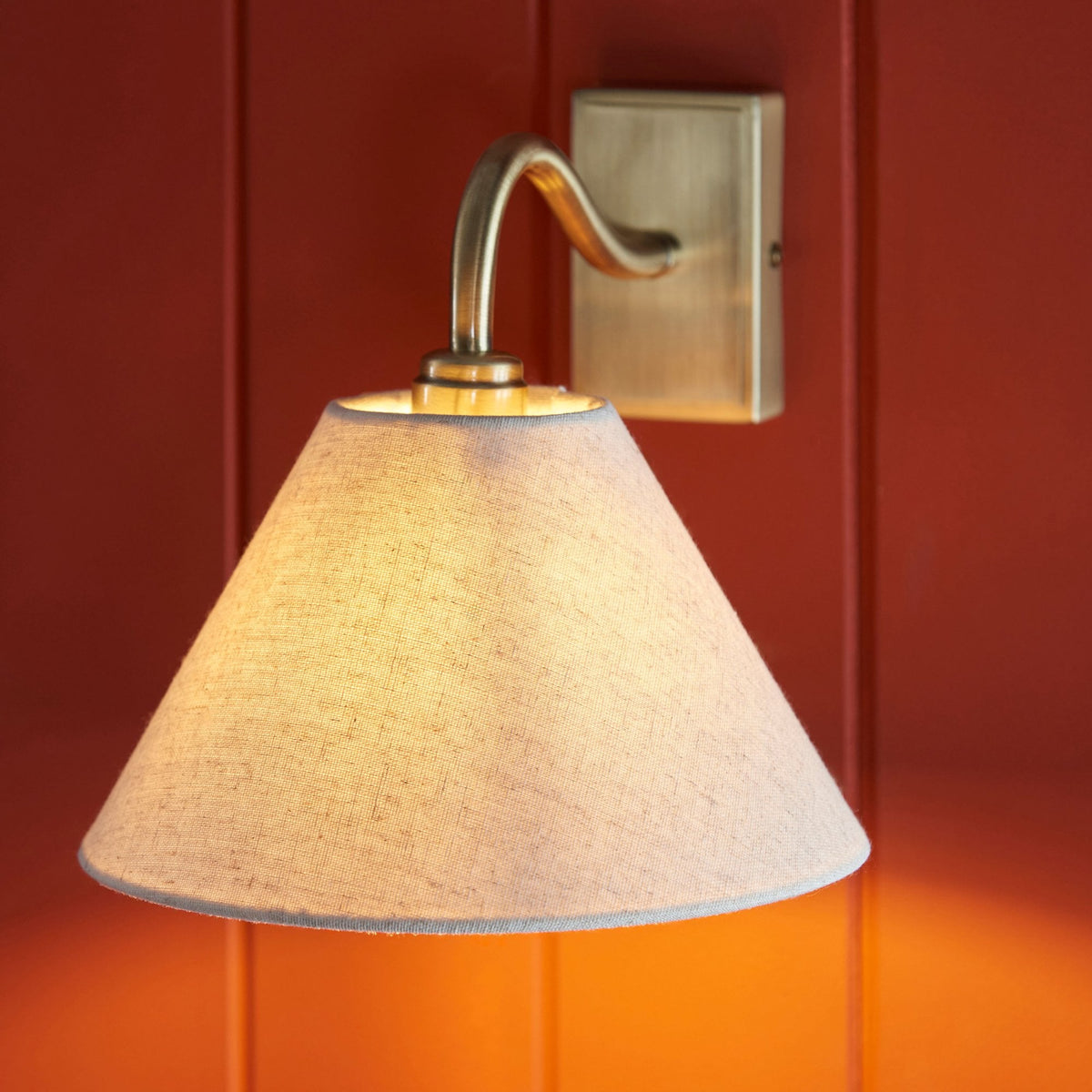 Aplique de pared de latón antiguo con pantalla cónica gris, iluminando una pared roja con vetas de madera verticales.