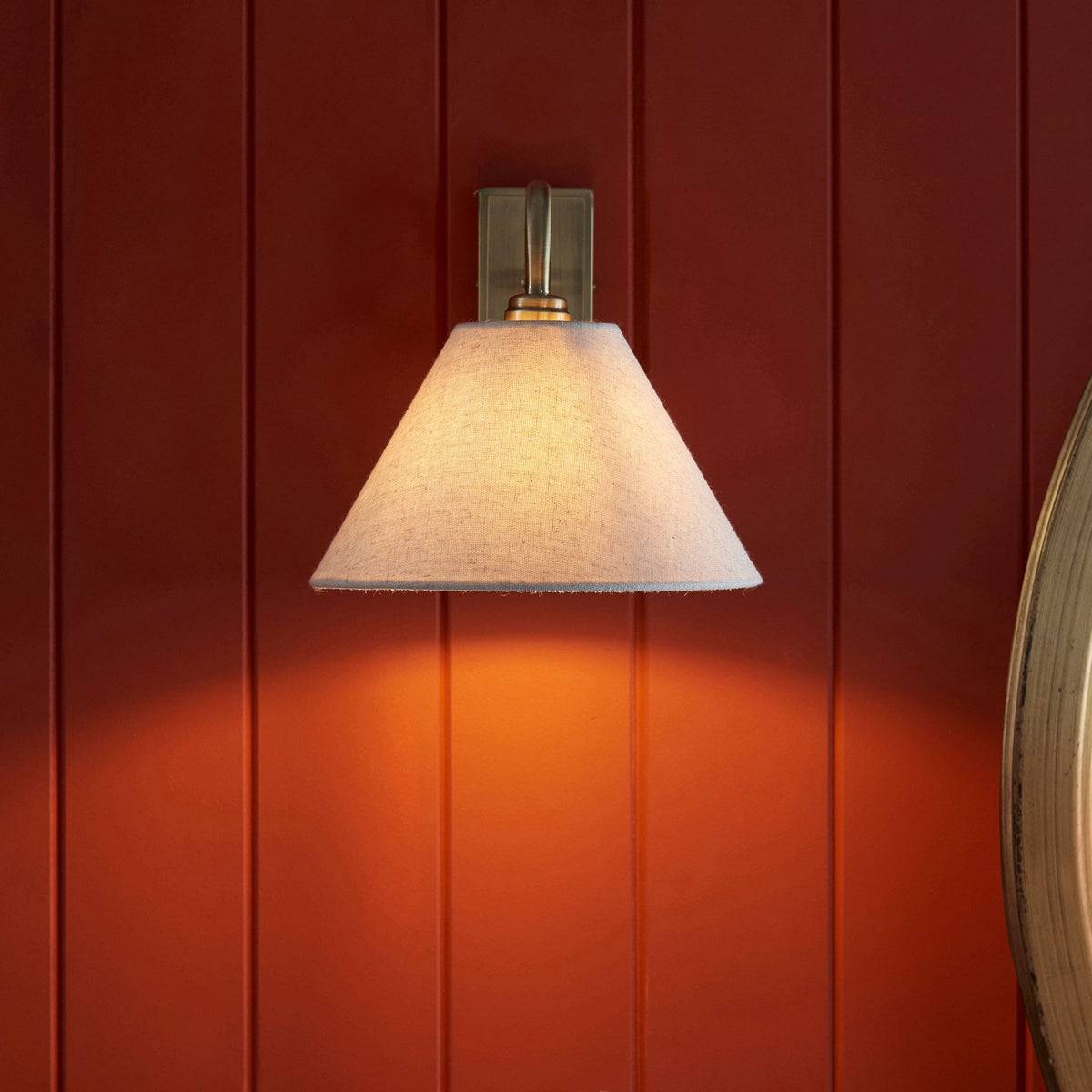 Aplique de pared de latón antiguo con pantalla cónica, brillando intensamente sobre un panel de pared de madera roja.