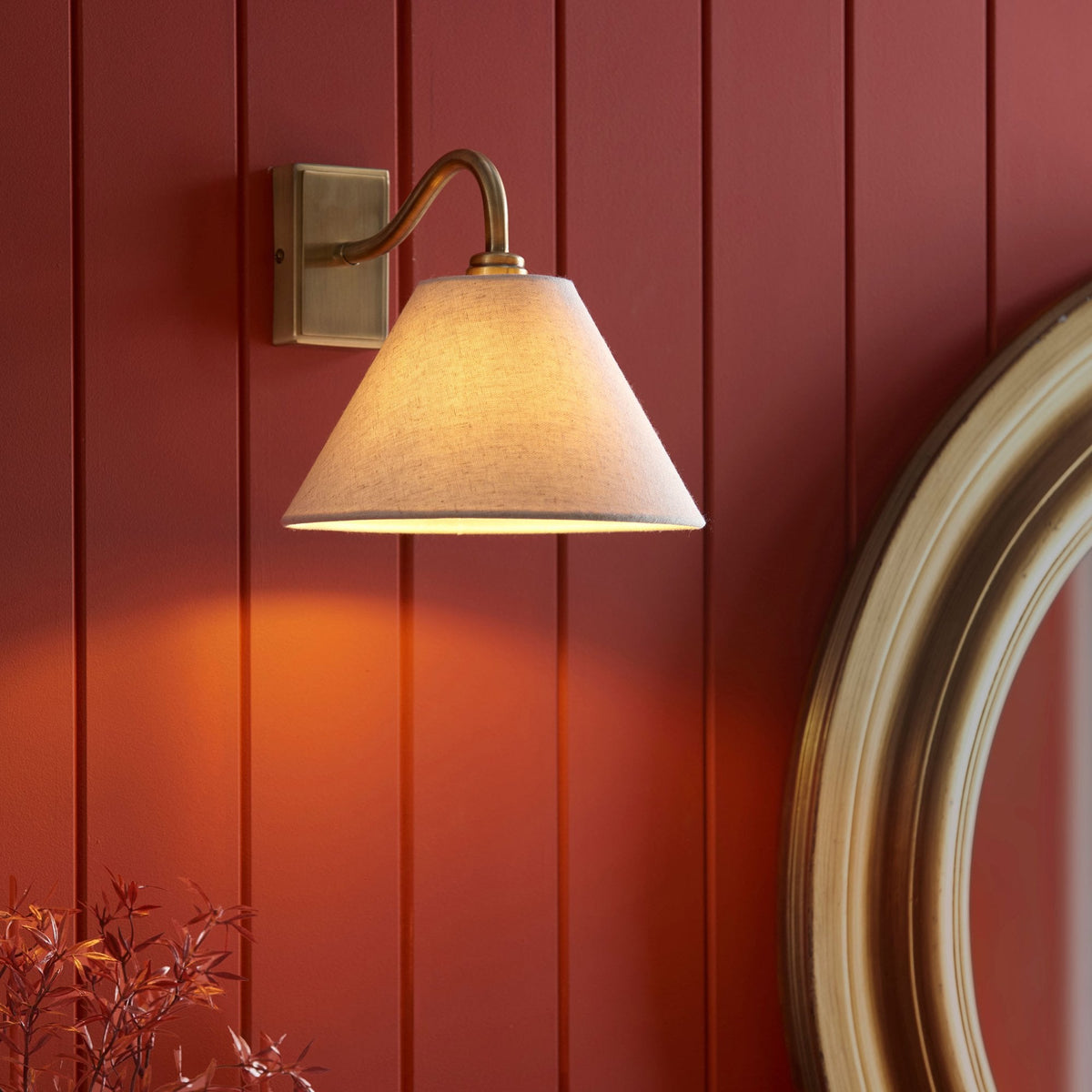 Aplique de pared de latón antiguo con pantalla cónica gris en una pared roja, con parte de un espejo visible.