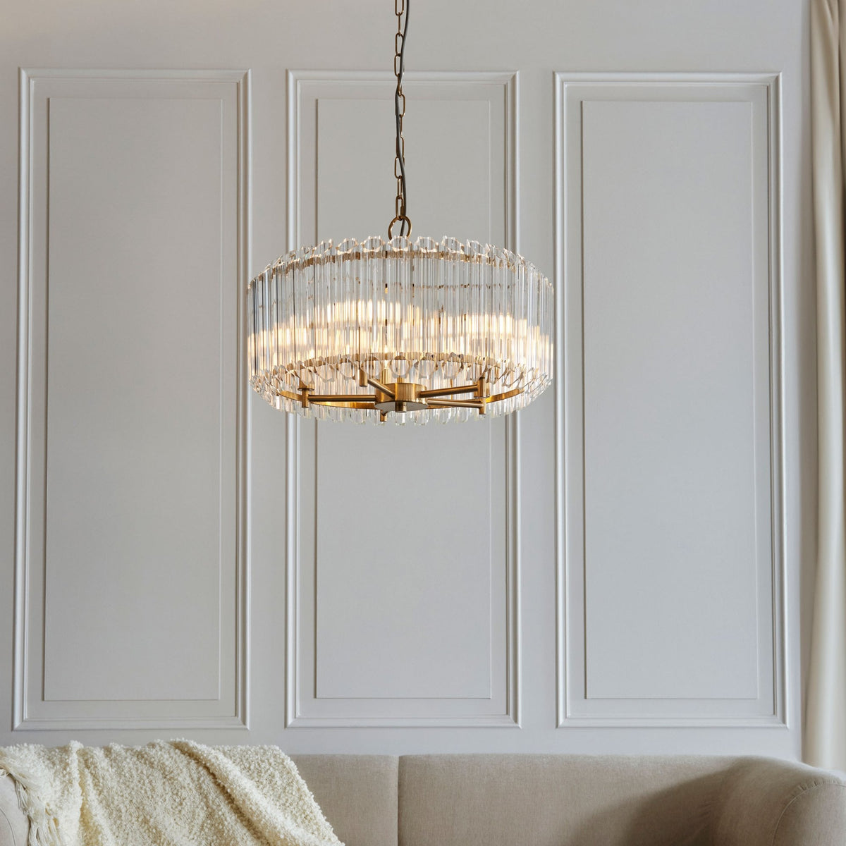 A golden chandelier with glass rods hanging in a room with white paneled walls and a beige sofa.