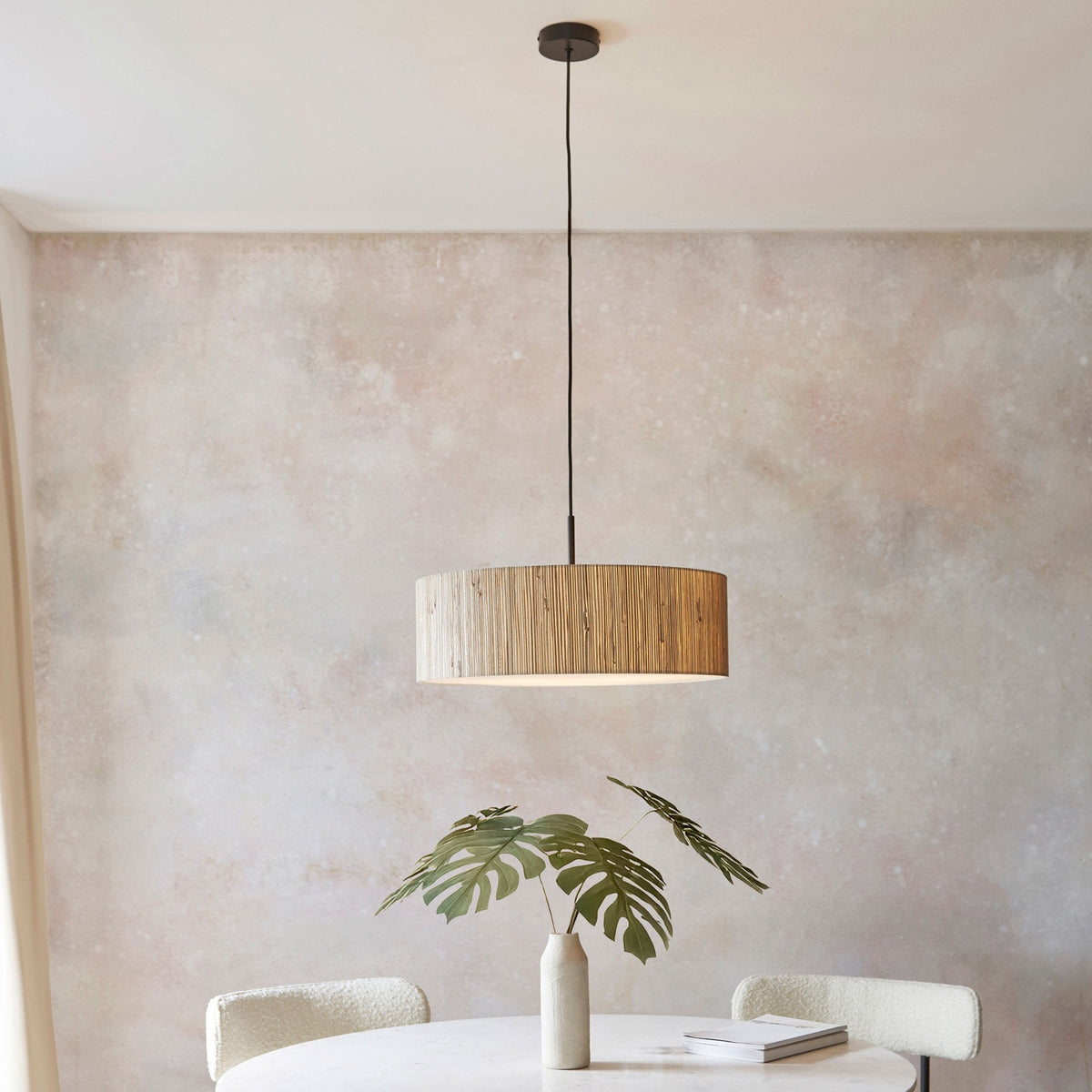 A wide shot of a dining room with a light wood slatted drum pendant light above a white table with a plant.