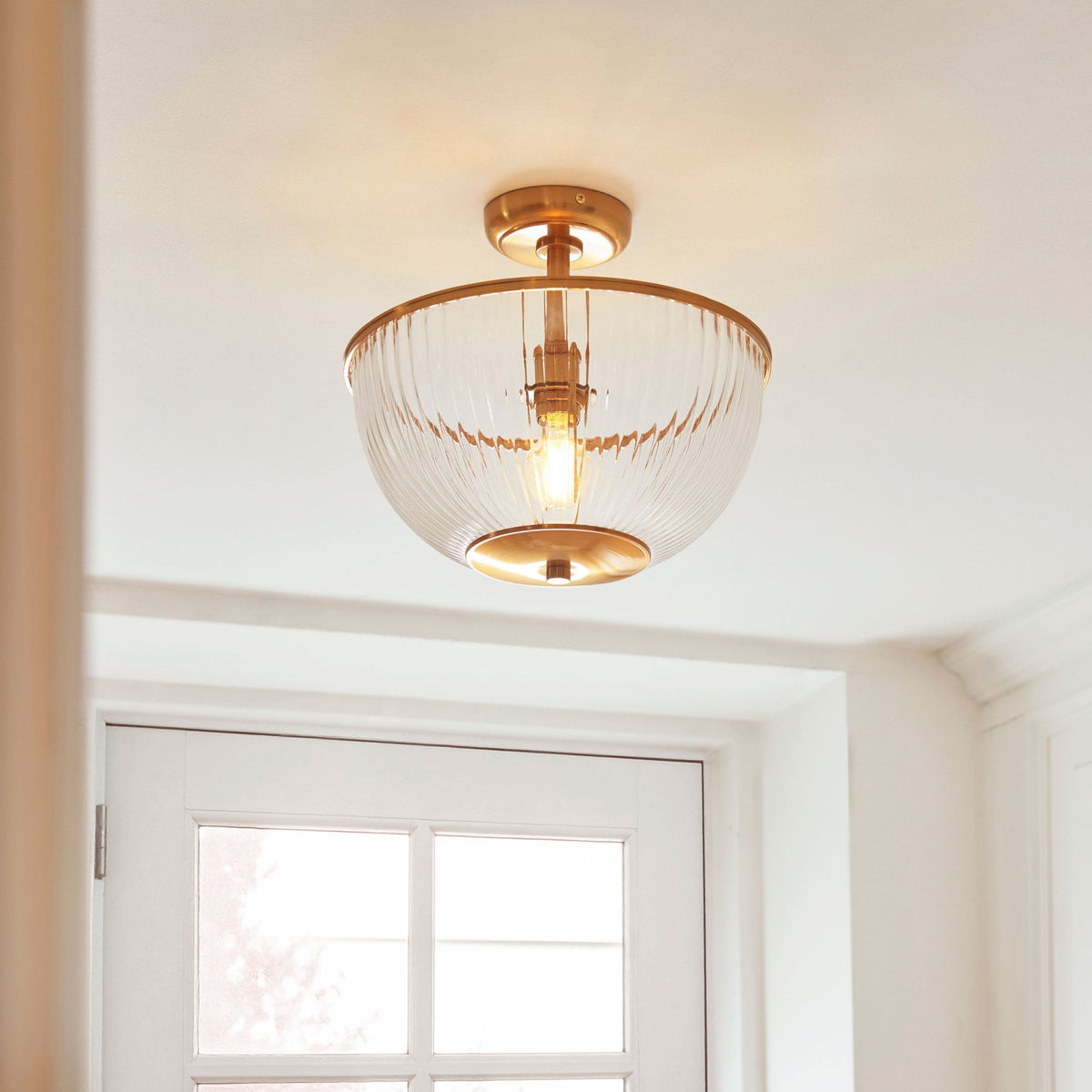 A semi-flush mount ceiling light with a ribbed glass shade and brass hardware hangs above a white door.