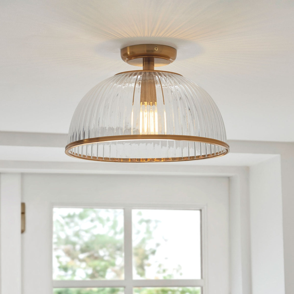 A clear, ribbed glass ceiling light with a brass base and trim, illuminating a room with a window.