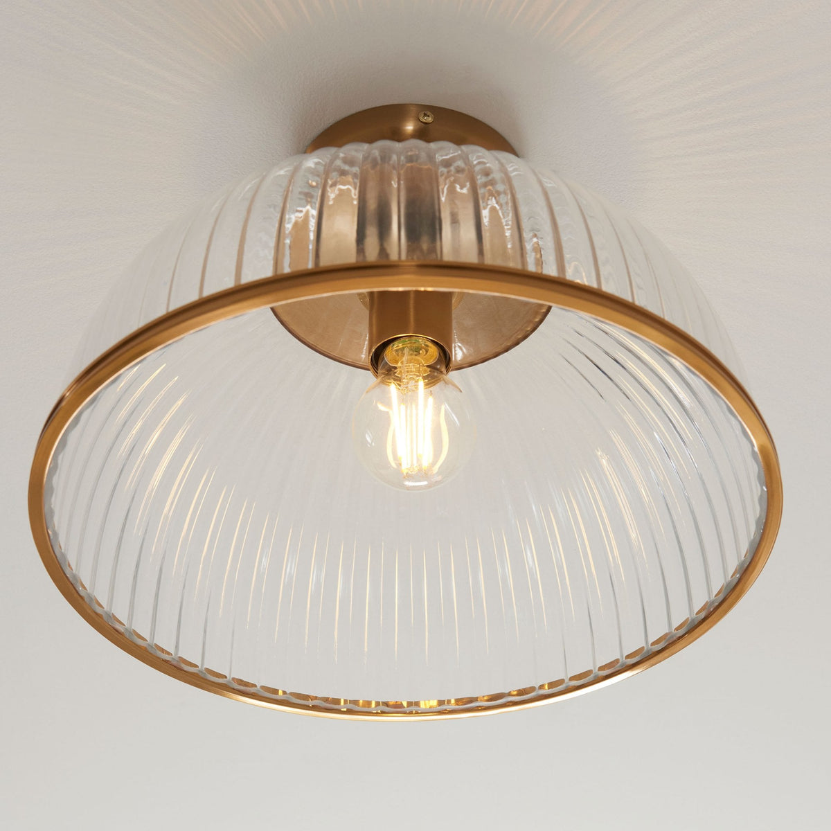 An upward-angled view of a clear, ribbed glass ceiling light with a brass base and trim, glowing warm.