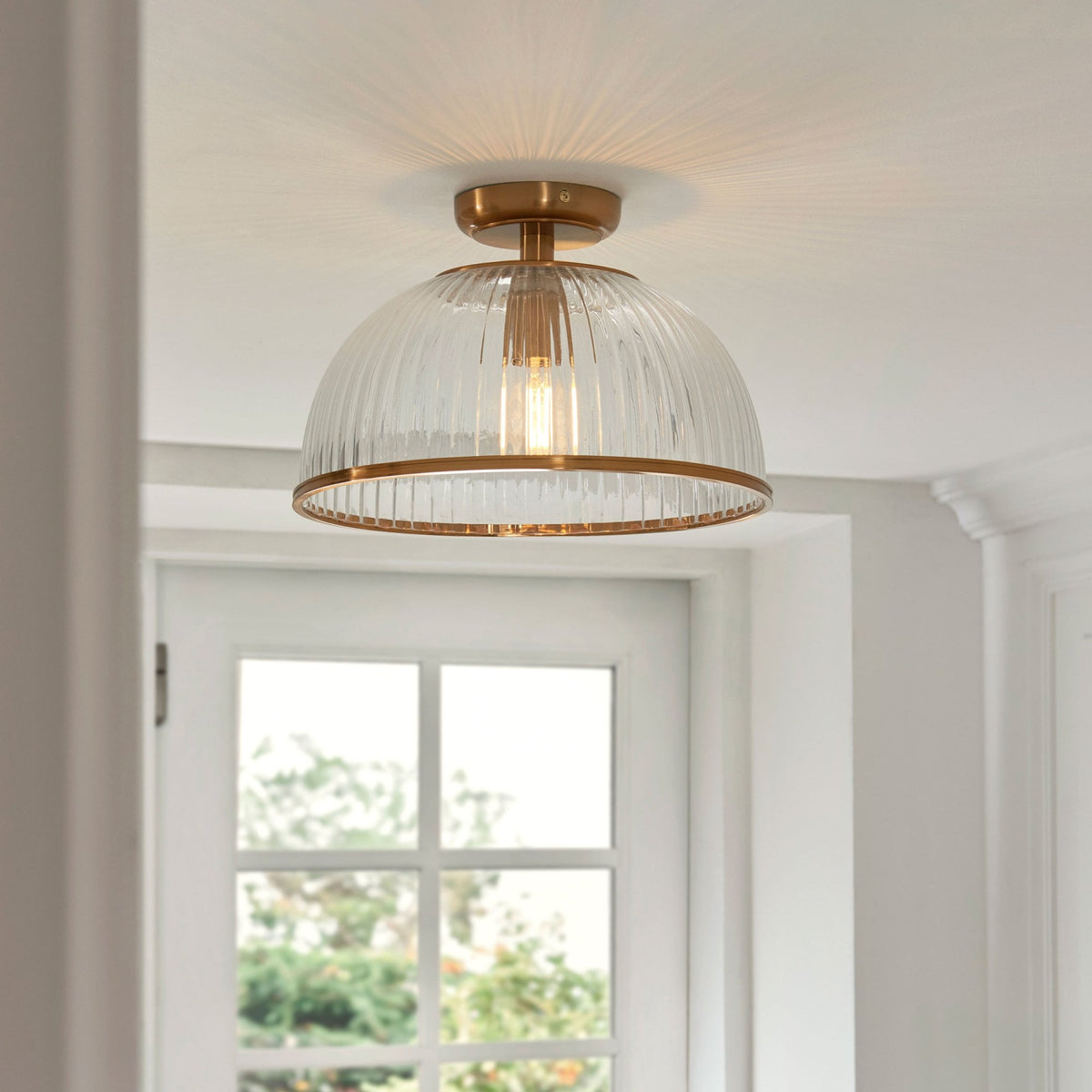 A clear, ribbed glass ceiling light with a brass base and trim, casting light above a window.