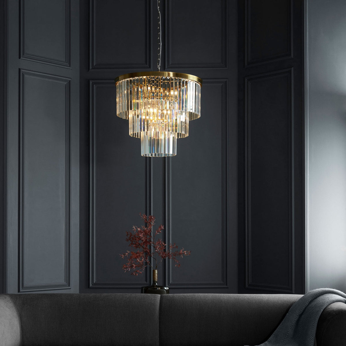 A tiered crystal chandelier illuminates a sophisticated room with dark paneled walls and a grey sofa.