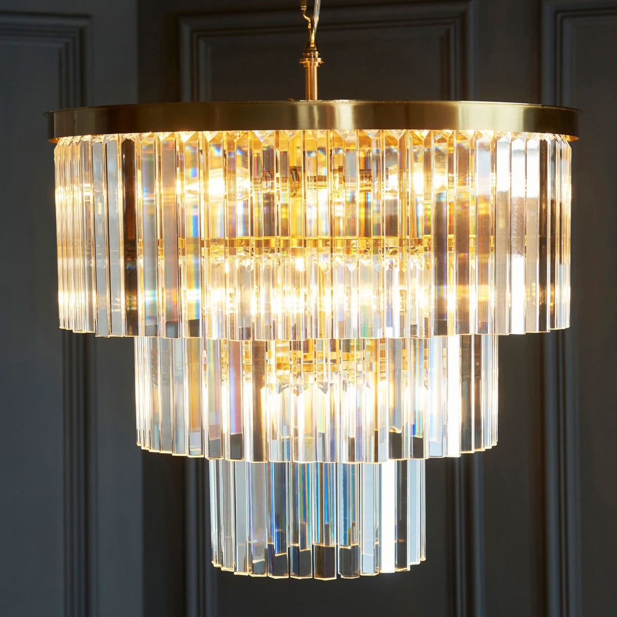 A dazzling, three-tiered crystal chandelier with a brass frame, casting a warm glow in a dark room.