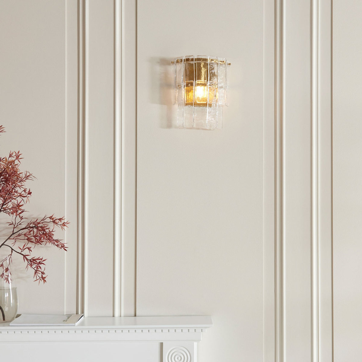 A sconce with textured glass and brass details mounted on a white wall above a fireplace mantel.
