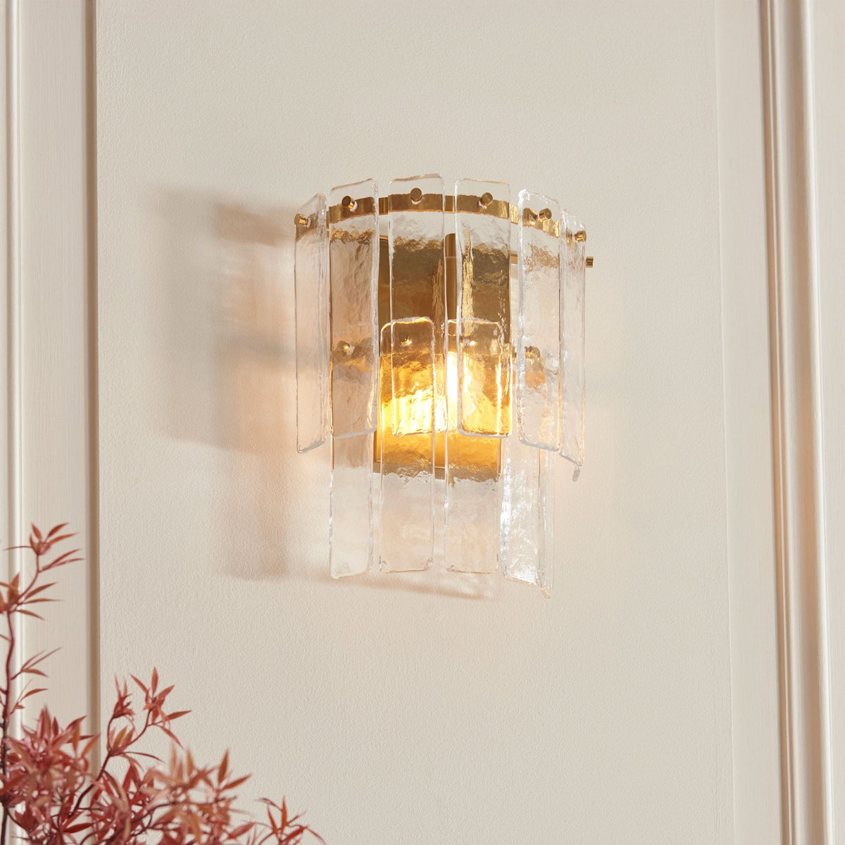 A sconce with textured glass panels and a brass frame casts a warm glow on a light-colored wall.