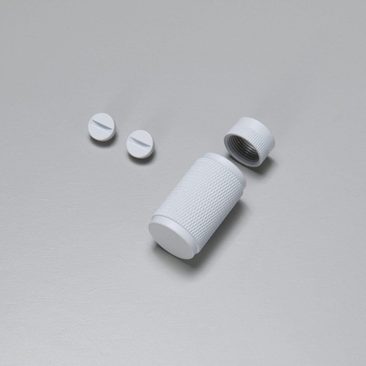 Two white pills and a white pill container with a textured grip, lid off, on a light gray surface.