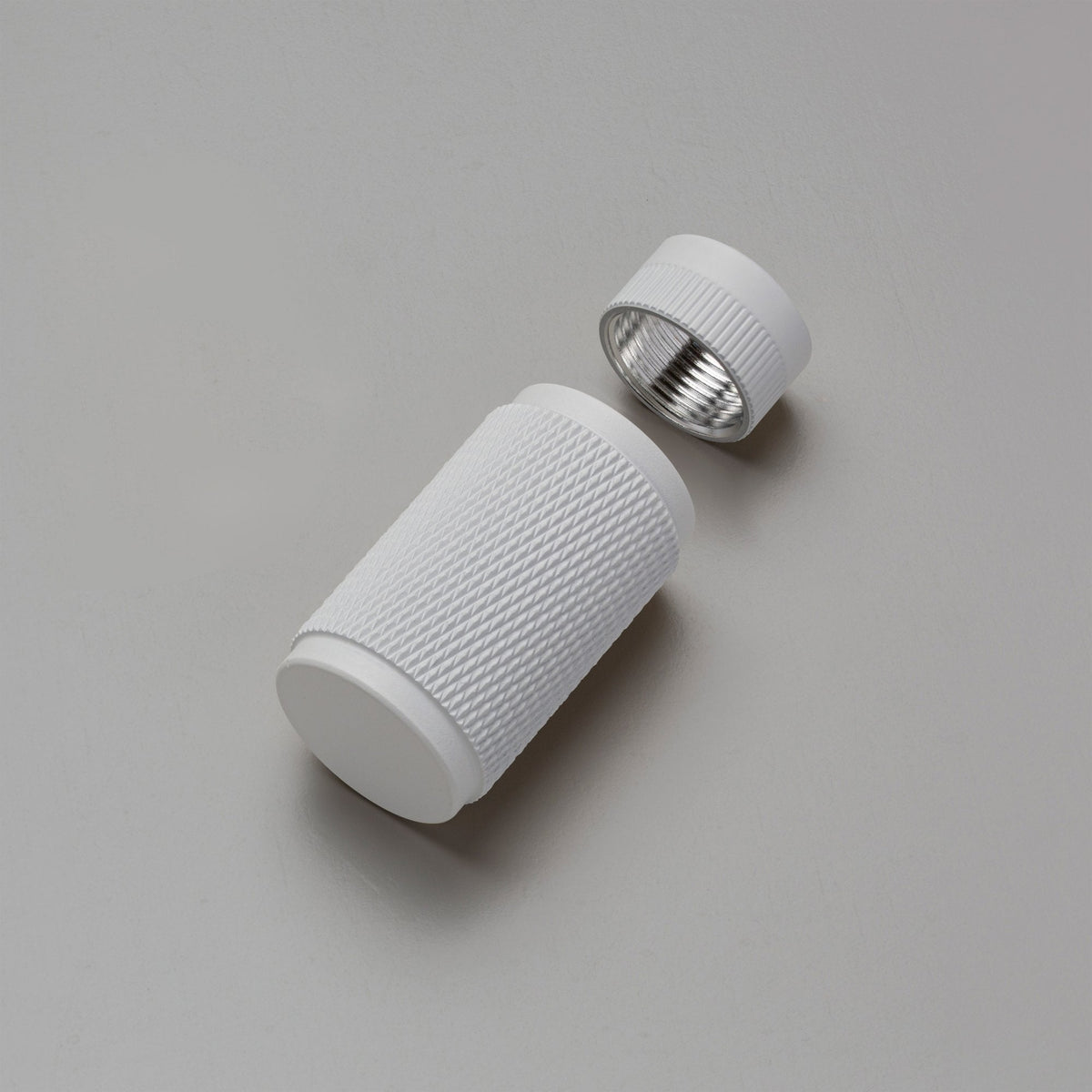 White cylindrical container with a textured grip, and its cap with a metallic interior, on a grey surface.