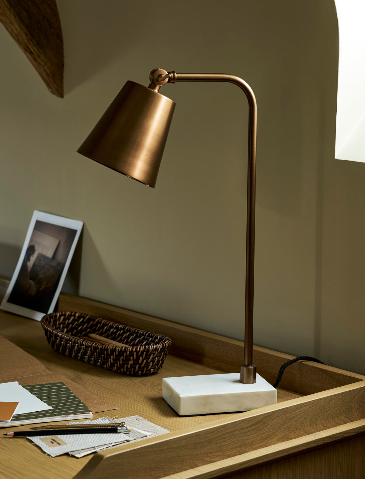 A modern table lamp with a warm bronze metal arm and shade, mounted on a white rectangular marble base, set on a wooden desk next to a woven basket and framed picture.