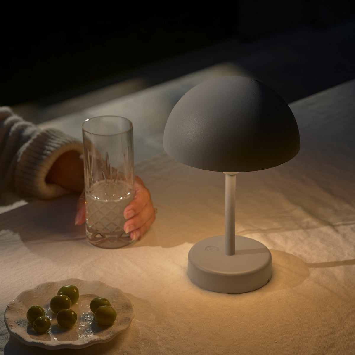 A person holding a glass of water next to a grey mushroom-shaped lamp and a plate of olives