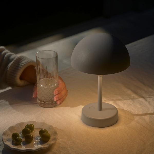 A person holding a glass of water next to a grey mushroom-shaped lamp and a plate of olives