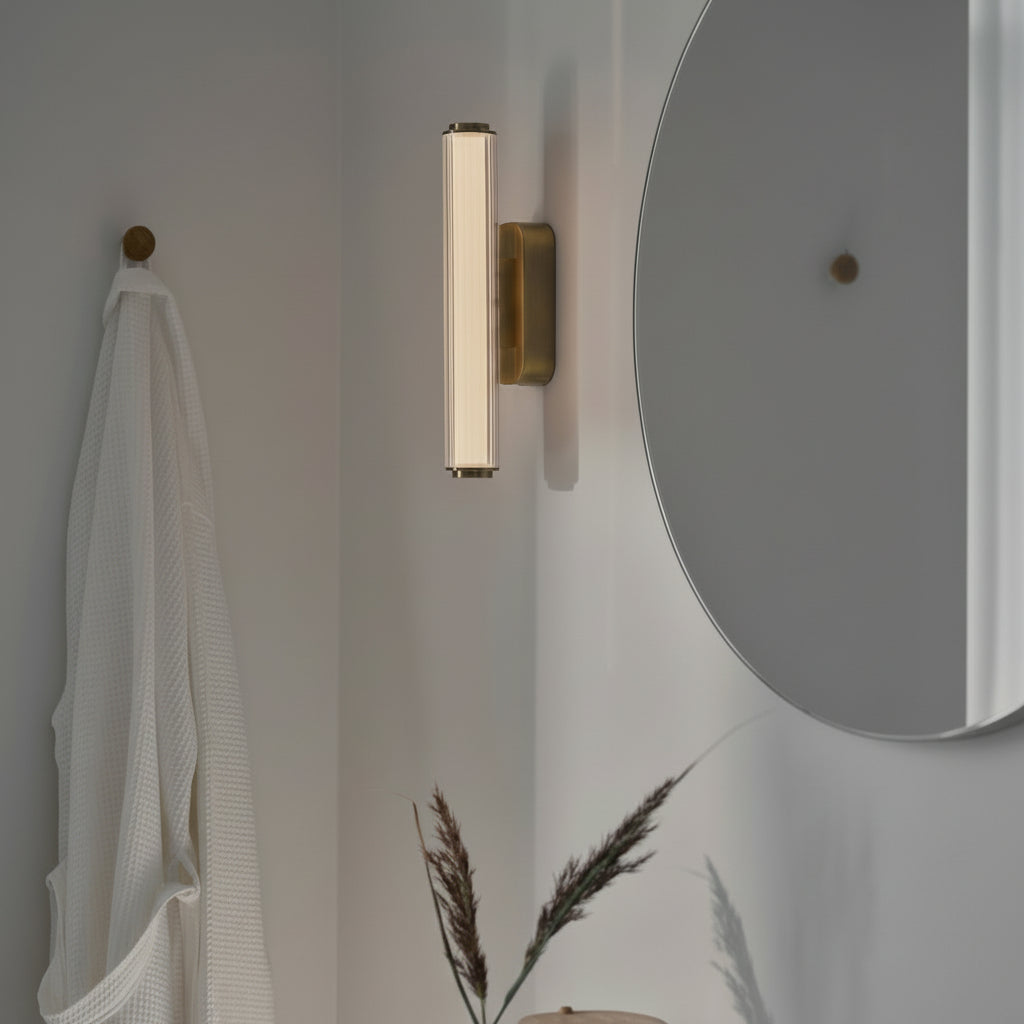 Vertical brass wall light with fluted glass shade next to a round mirror and white robe