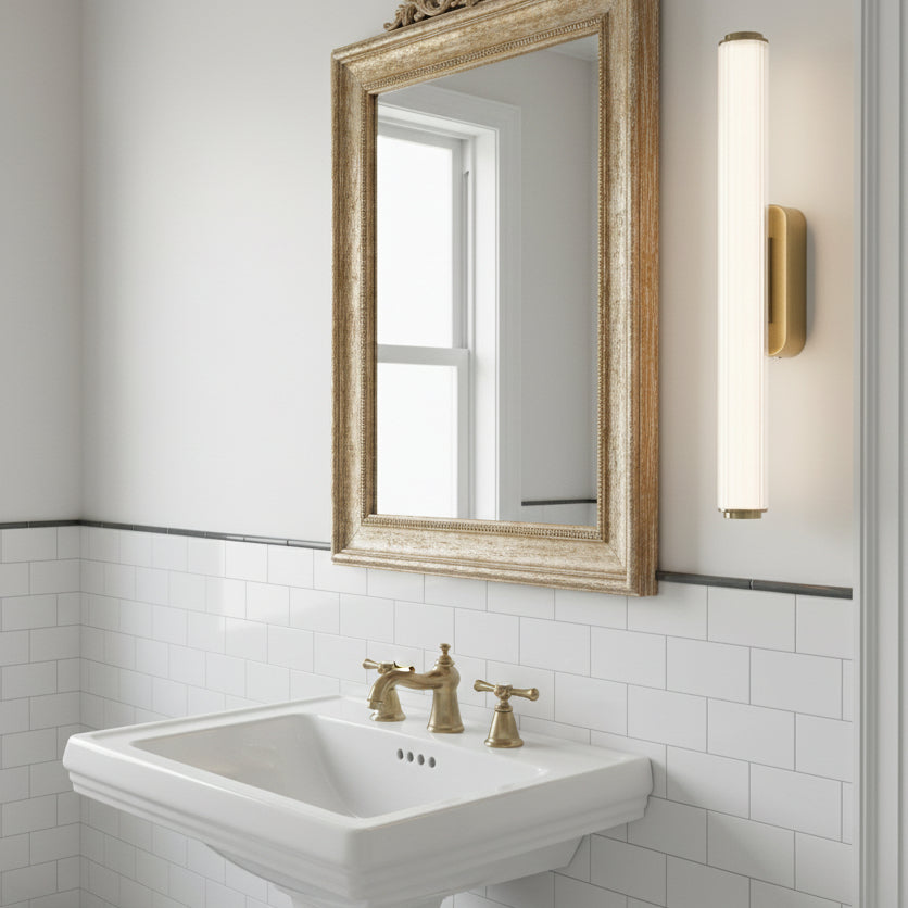 Bathroom with white subway tiles, a pedestal sink with brass faucet, a gold framed mirror, and a wall sconce