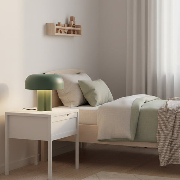A bedroom with a bed, a nightstand, and a shelf. The bed has a light green duvet and pillows. The nightstand has a green lamp. The shelf has wooden blocks.