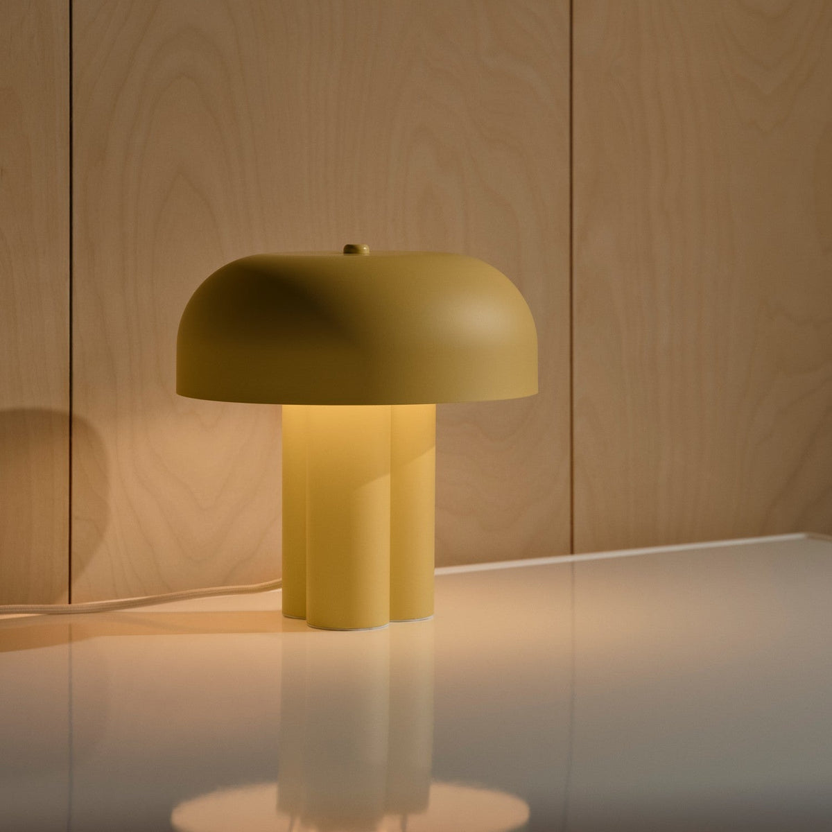 Yellow table lamp with a mushroom-shaped shade and three cylindrical legs, casting a warm glow on a white surface