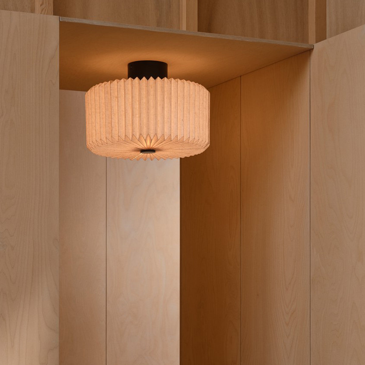 A round, pleated, light-colored ceiling lamp illuminates a corner of a room with light wood paneling