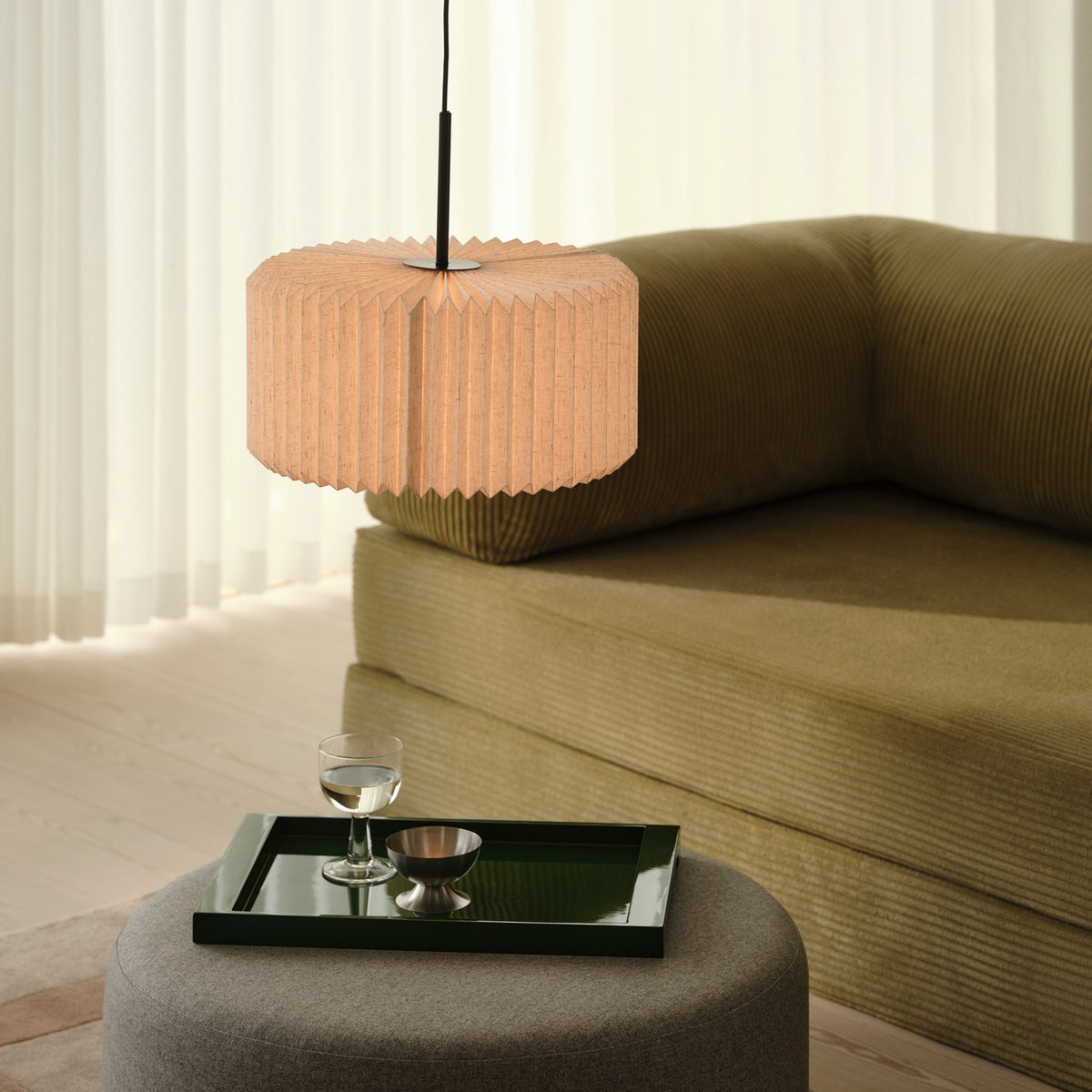 Pleated pendant lamp over a grey ottoman with a green tray, glass, and metal bowl