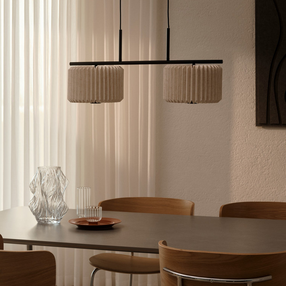 Two pleated fabric pendant lights hang above a dining table with a glass vase and wooden chairs