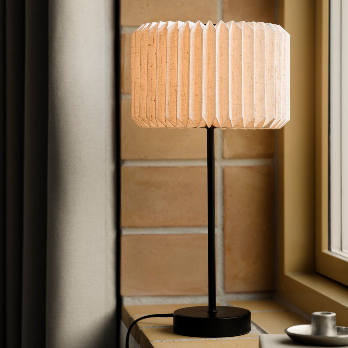 A table lamp with a pleated fabric shade and a black base sits on a windowsill next to a window