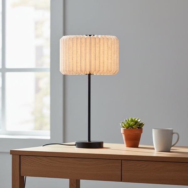 Table lamp with a pleated fabric shade, a black base and stem, on a wooden desk with a plant and mug