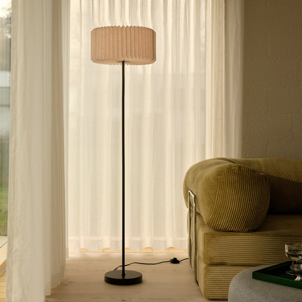 Tall floor lamp with a pleated fabric shade next to a corduroy sofa and sheer curtains