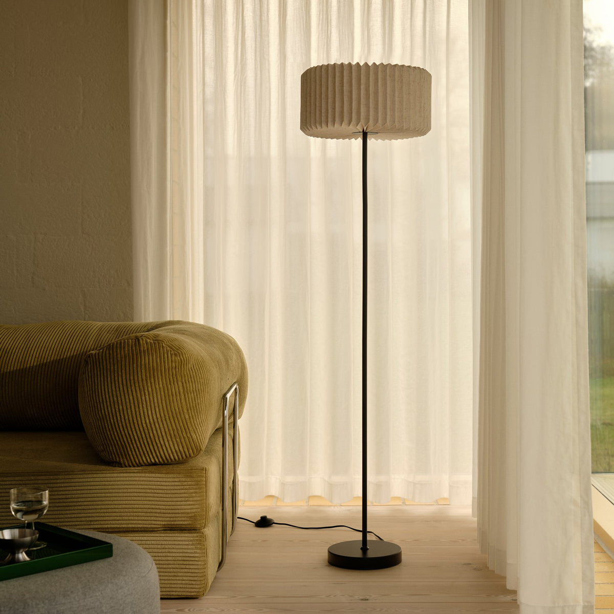 Tall floor lamp with a pleated shade next to a corduroy sofa and sheer curtains