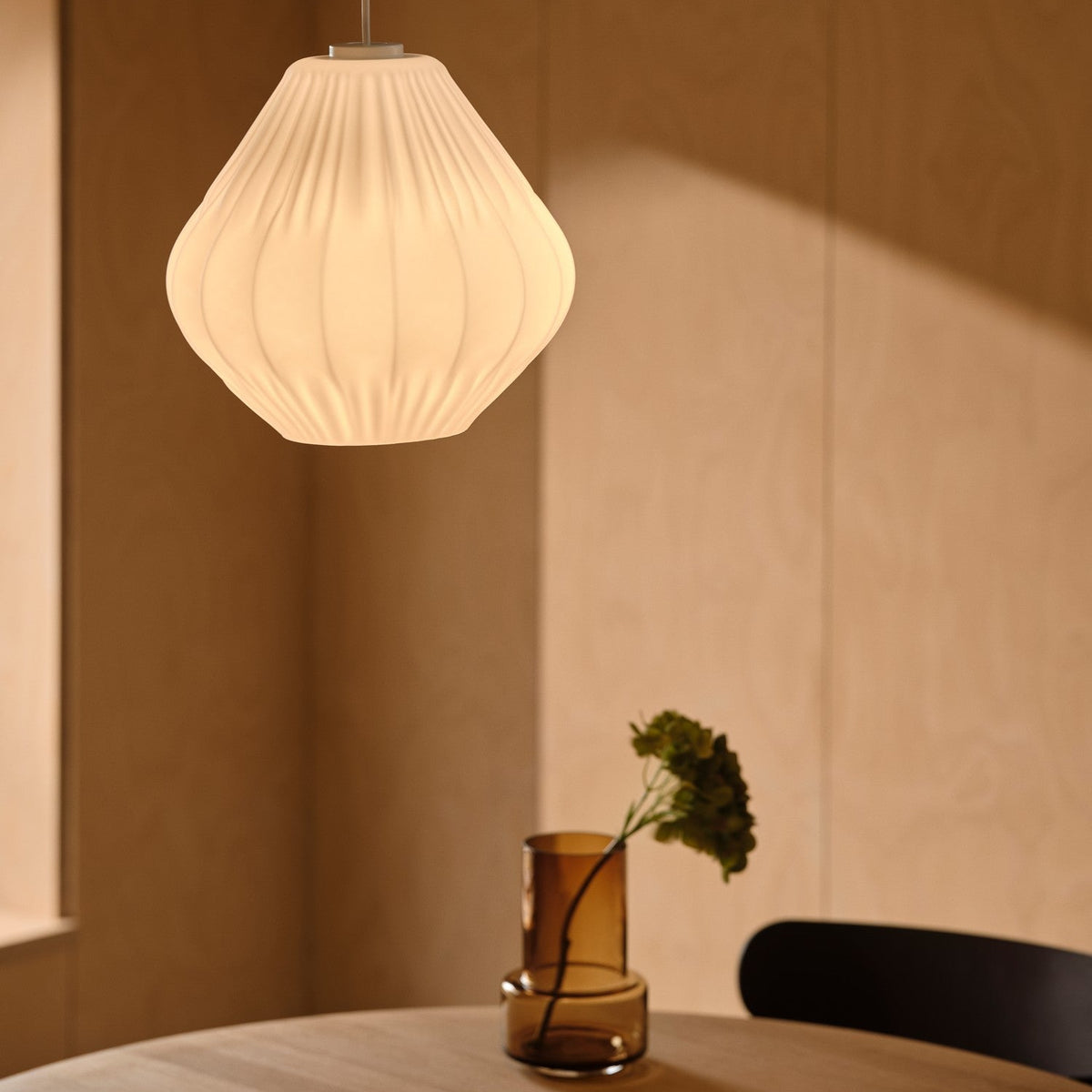 White pleated pendant light above a wooden table with a brown vase and green plant