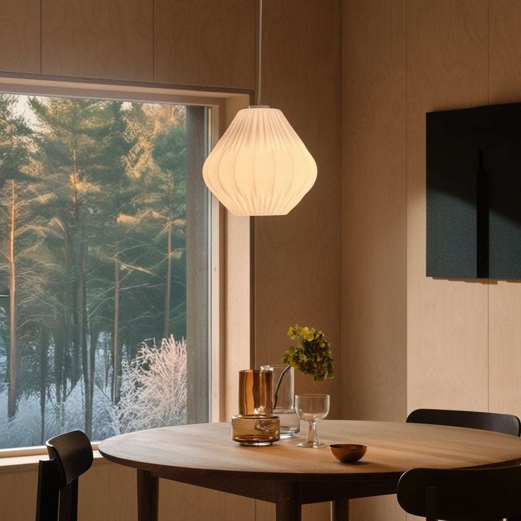 A white pleated pendant light hangs above a round wooden table with vases and glasses, next to a window