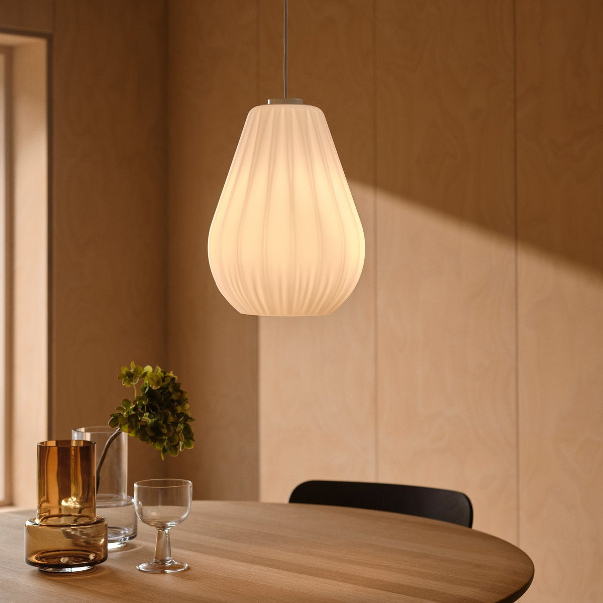 White pleated pendant lamp hanging over a light wood round table with glass vases and a wine glass