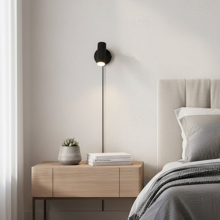 Black wall light with a ribbed design and a visible cord, illuminating a bedside table with a plant and books