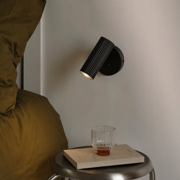 Black fluted wall light with warm glow above a bedside table with a book and glass