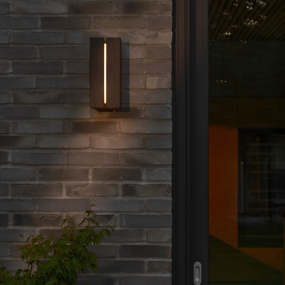 Black outdoor wall light with a vertical light strip on a dark brick wall next to a glass door